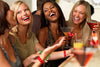 Four young women laughing and holding cocktails at a bar, with one woman wearing a bright red wristband.